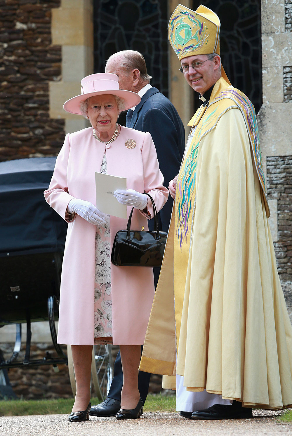 Her Majesty, Queen Elizabeth II wears a pale pink jacquard wool coat worn over a floral silk dress with a matching pale pink hat and her signature patent leather black purse and heels.