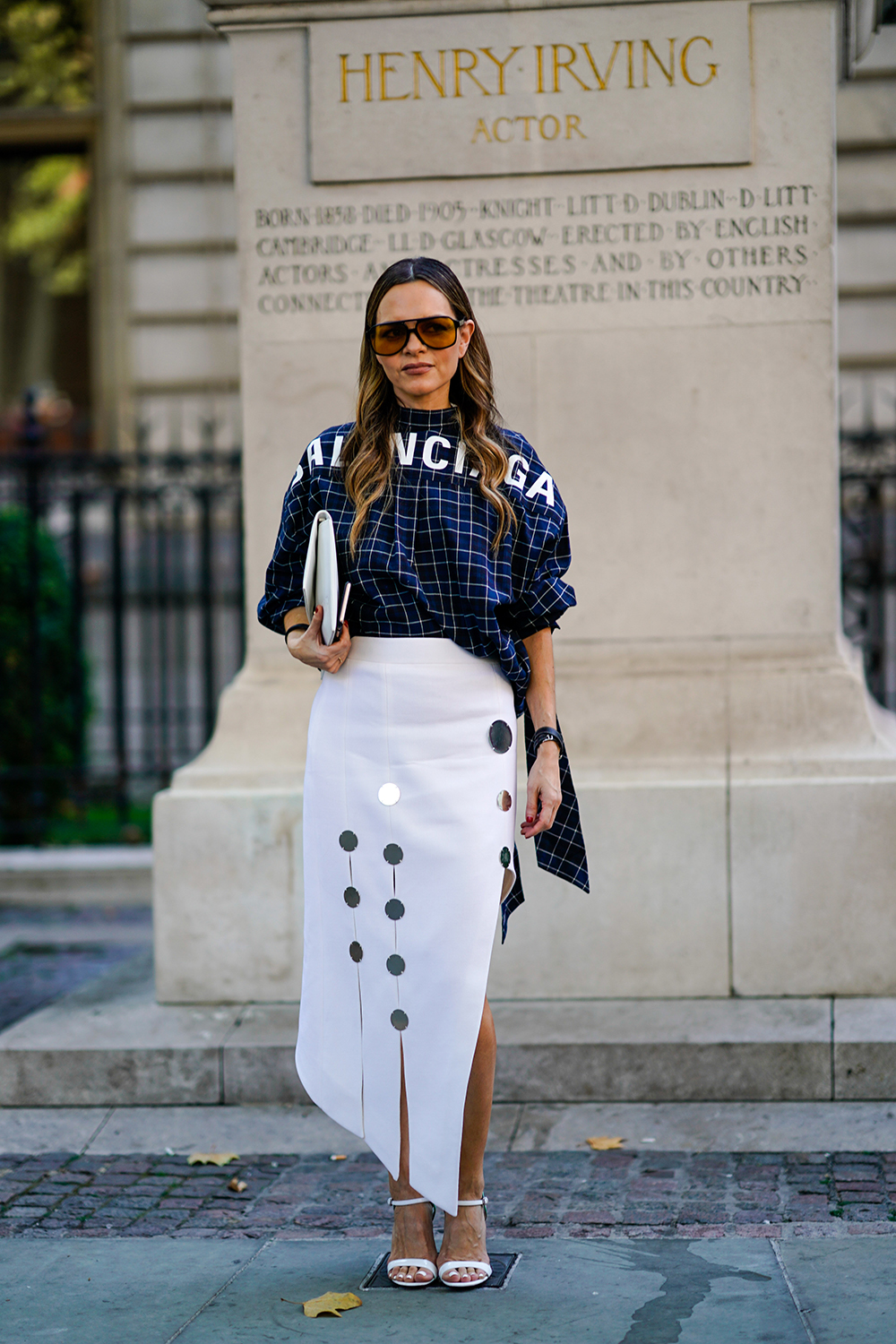 A guest wears sunglasses, a a Balenciaga blue checked top, white skirt, a white clutch, during London Fashion Week September 2018 on September 17, 2018 in London, England.