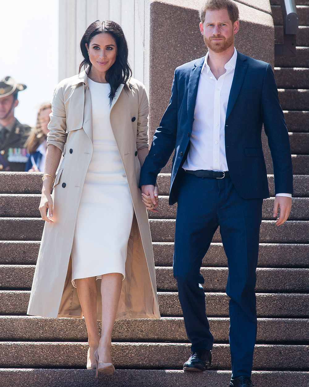October 16, 2018: Meghan, Duchess of Sussex descends the stairs outside the Sydney Opera House on day one of her and husband Prince Harry, Duke of Sussex' tour of Australia. Meghan wears a white sleeveless sheath dress aptly named Blessed by Australian designer Karen Gee underneath a camel trench coat. Meghan accessorised with nude suede pumps by Stuart Weitzman and butterfly earrings and matching bracelet that belonged to the late Princess Diana.